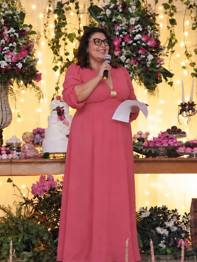 Carla Camilo de vestido rosa, ao microfone, diante de uma mesa de doces com flores e velas