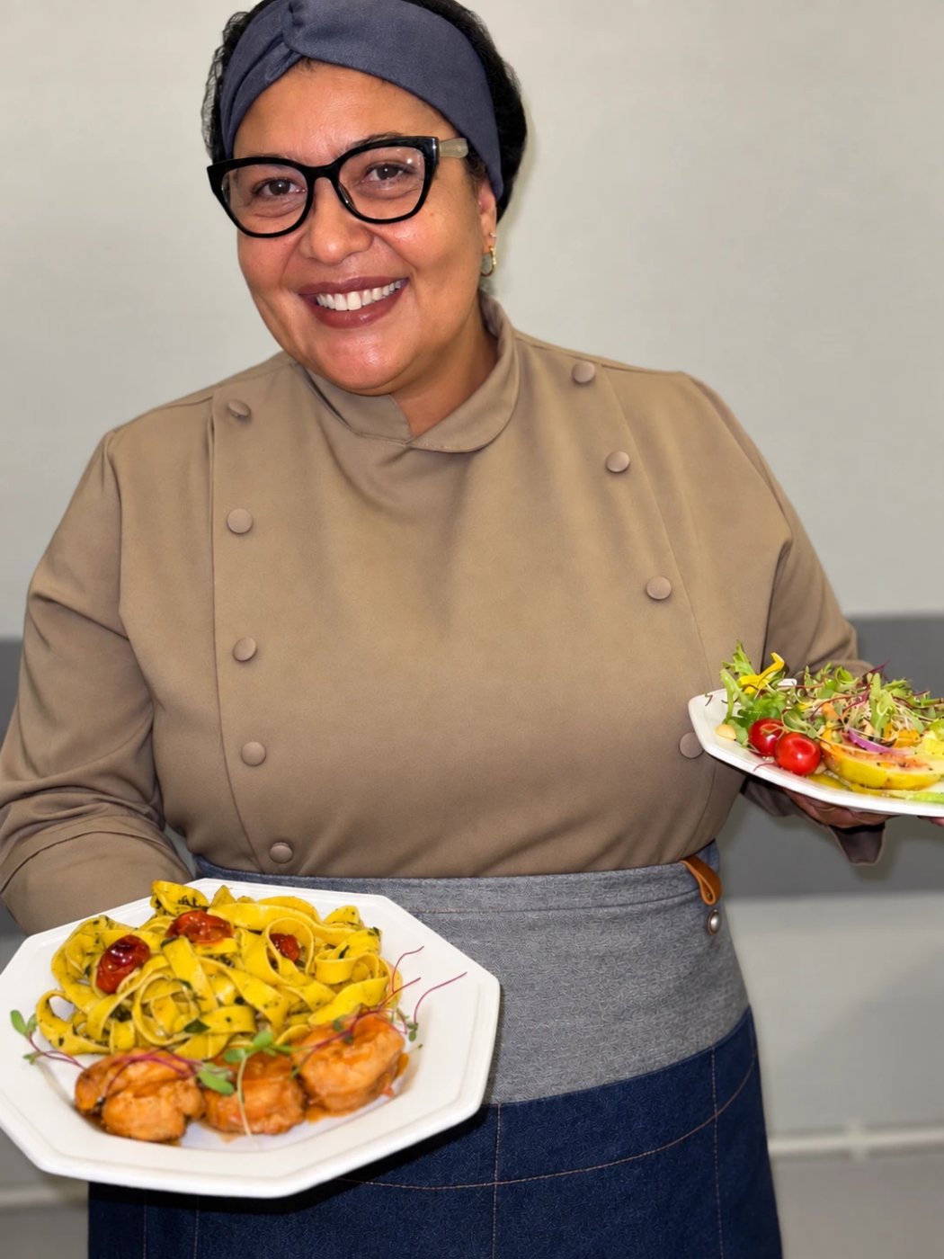 Carla Camilo de dólmã, sorrindo, com um tagliatelle ao pesto e uma salada com flores nas mãos