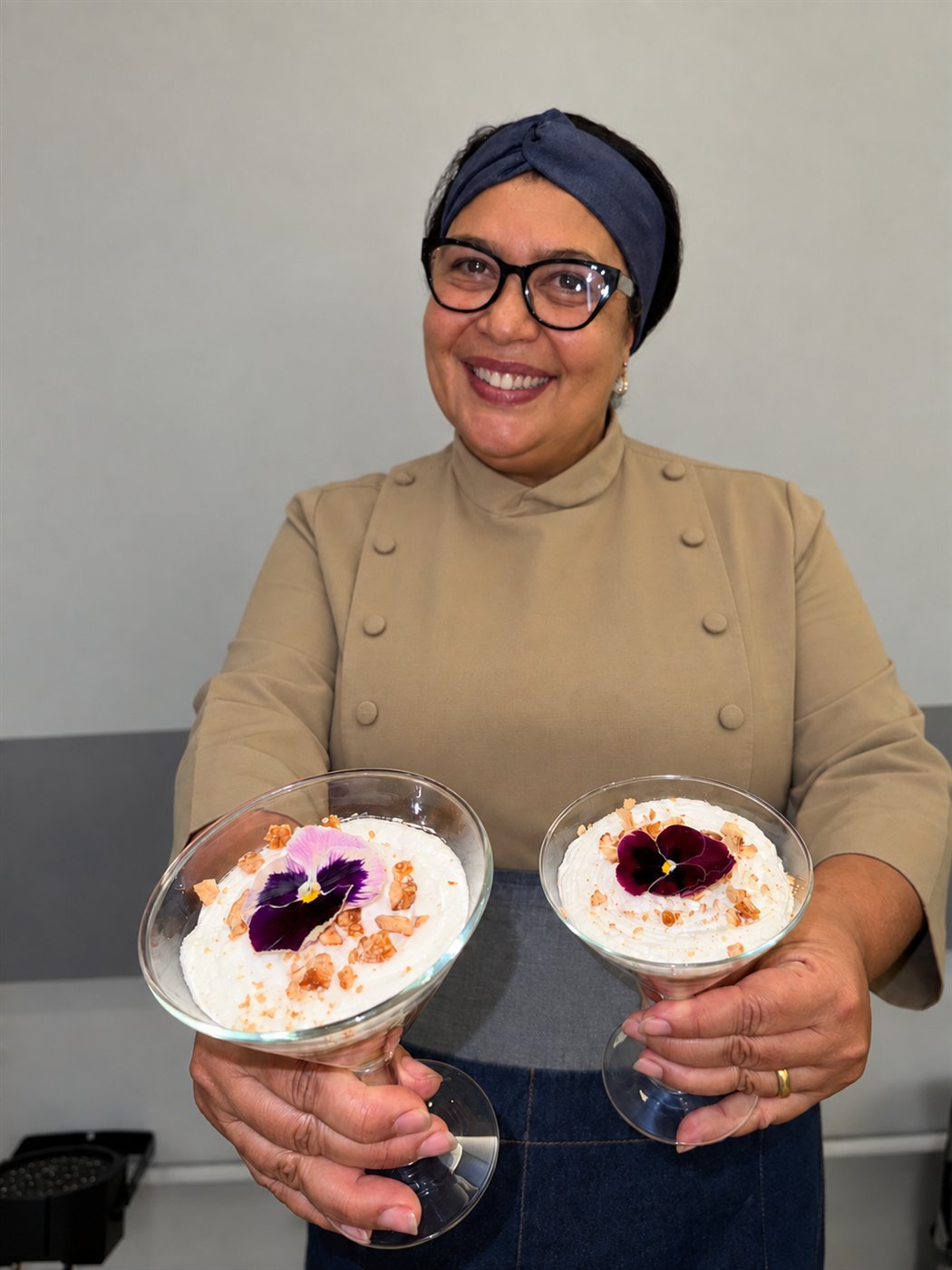 Carla Camilo sorrindo, oferecendo duas taças de sobremesa com flor amor-perfeito